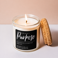 A lit jar candle with a black label reading 'PAIN HAS A Purpose' and 'Hand Poured Coconut Wax with Juice Tea' on a clean white surface against a soft beige background. A cork stopper leans against the jar.