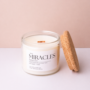 A jar candle labeled 'His Miracles' with white wax and a wooden wick, shown next to its cork lid on a clean white surface against a soft blush pink background.