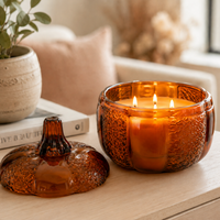 An amber glass candle holder with a matching lid sits on a light wood table beside a lit amber candle. The scene is styled in a cozy indoor setting with a potted plant, books, and soft background decor.
