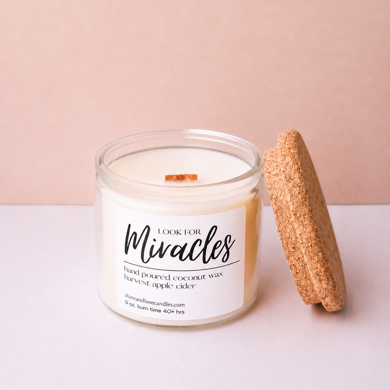 A clear glass jar candle with white wax and a wooden wick, labeled 'Cook For Miracles Harvest Apple cider,' shown next to a cork lid on a pale pink background.