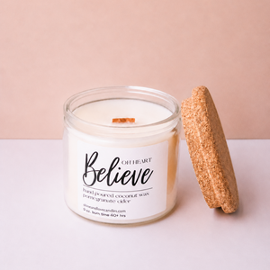 A white soy wax candle in a clear glass jar with a white label reading 'Believe' and 'Of Heart', shown next to a round cork stopper on a light pink and white surface.