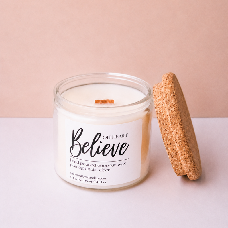 A white soy wax candle in a clear glass jar with a white label reading 'Believe' and 'Of Heart', shown next to a round cork stopper on a light pink and white surface.