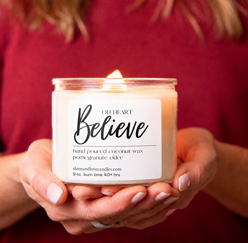 A person in a burgundy top holds a lit candle in a clear glass jar. The candle has a white label that reads 'OH HEART Believe' and 'hand poured coconut wax pomadeurate cider.