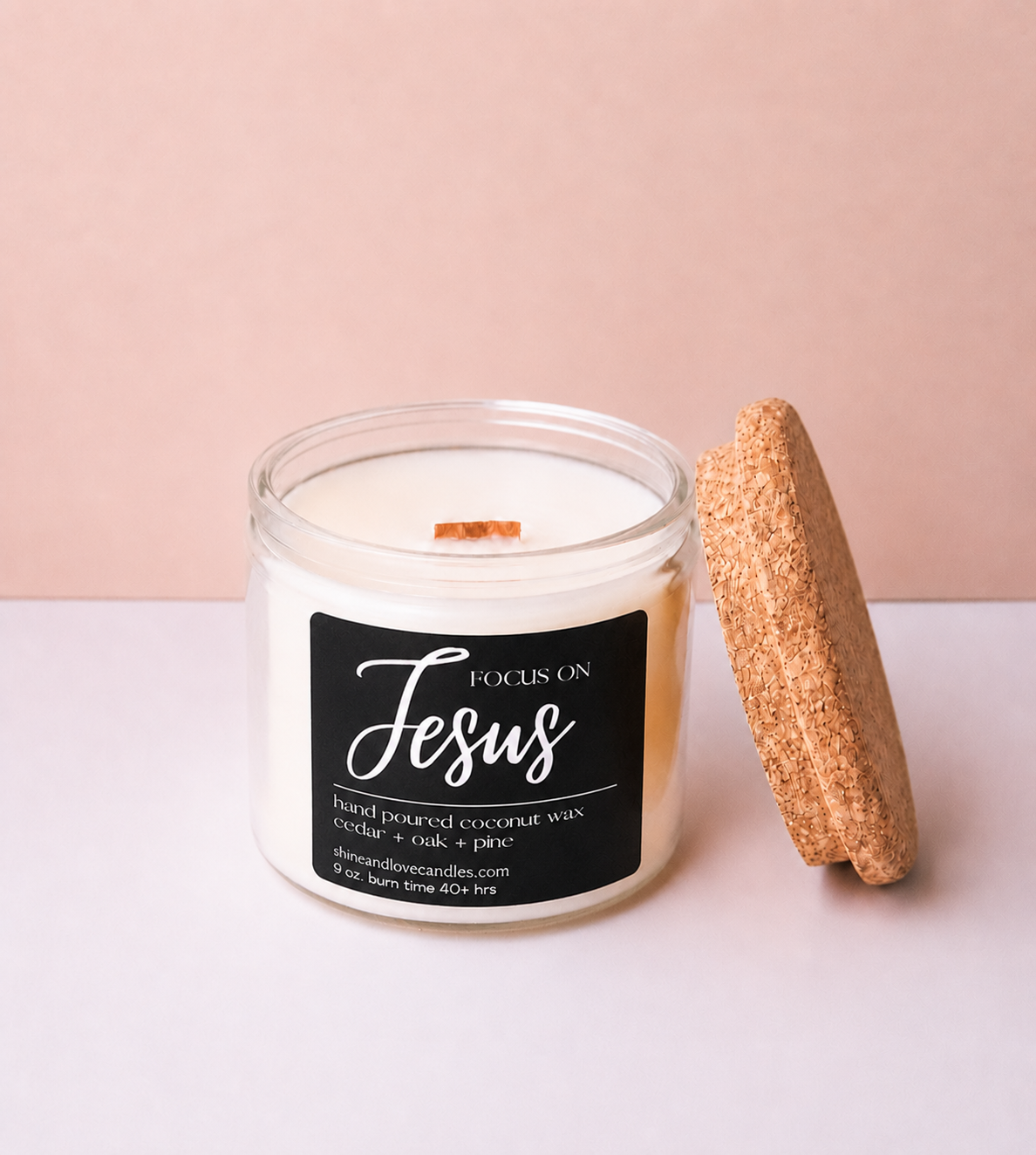 A clear glass jar candle with white wax and a black label reading 'Focus on Jesus' in white script, shown next to a cork stopper on a pale pink and white background.