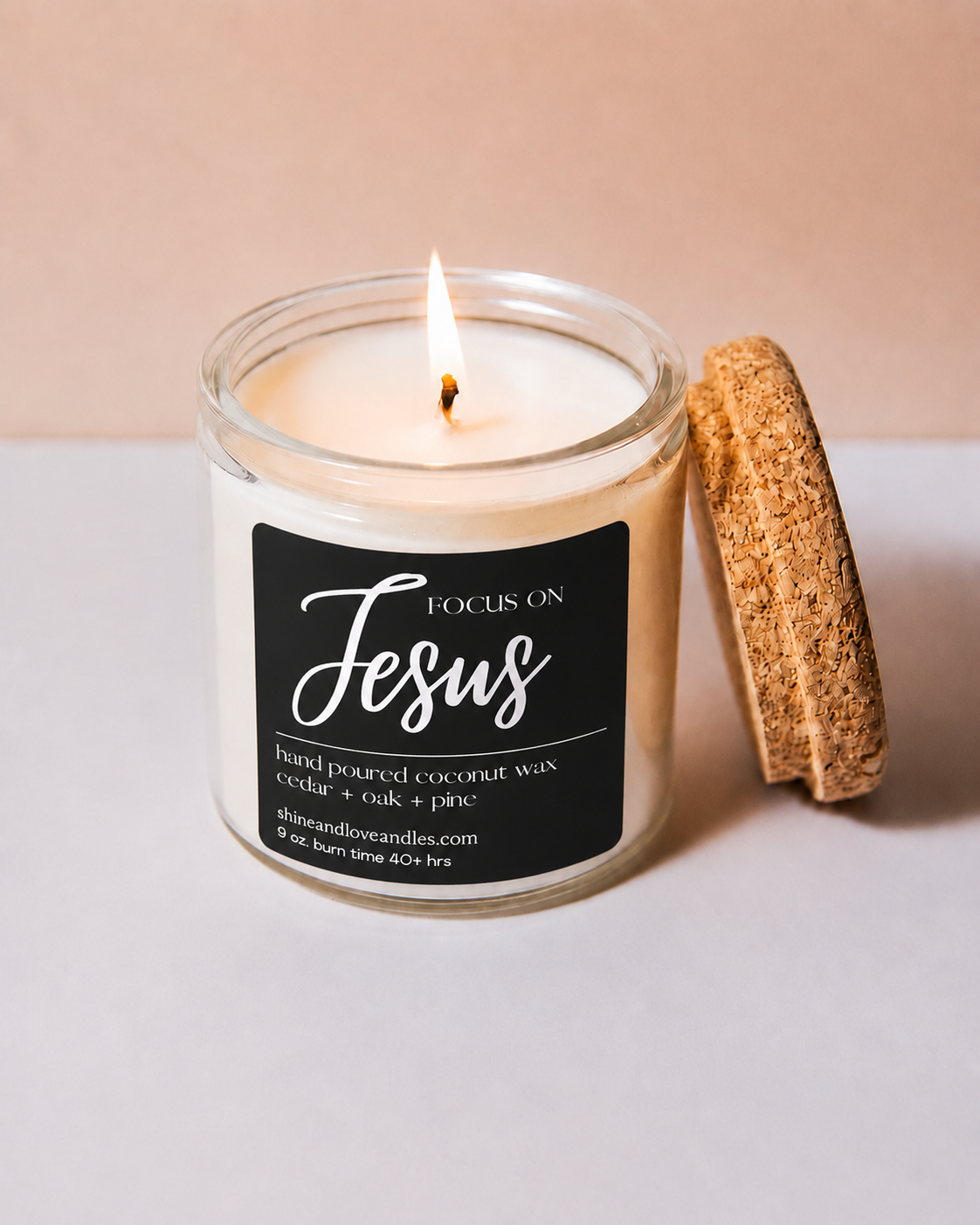 A lit candle in a clear glass jar with a black label reading 'Focus on Jesus' and 'Hand poured coconut wax cedar + oak + pine.' A cork lid leans against the jar on a light surface against a warm beige background.