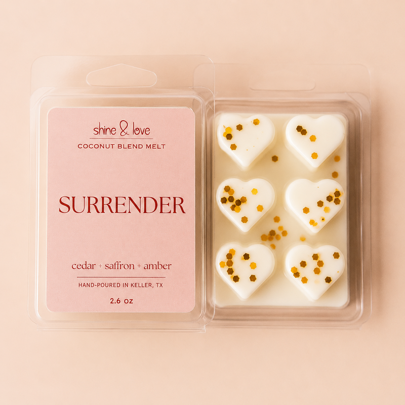 A 2.6 oz clamshell package of 'Surrender' coconut blend melt wax by shine & love, featuring six white heart-shaped wax melts decorated with gold glitter, set against a solid pale pink background.